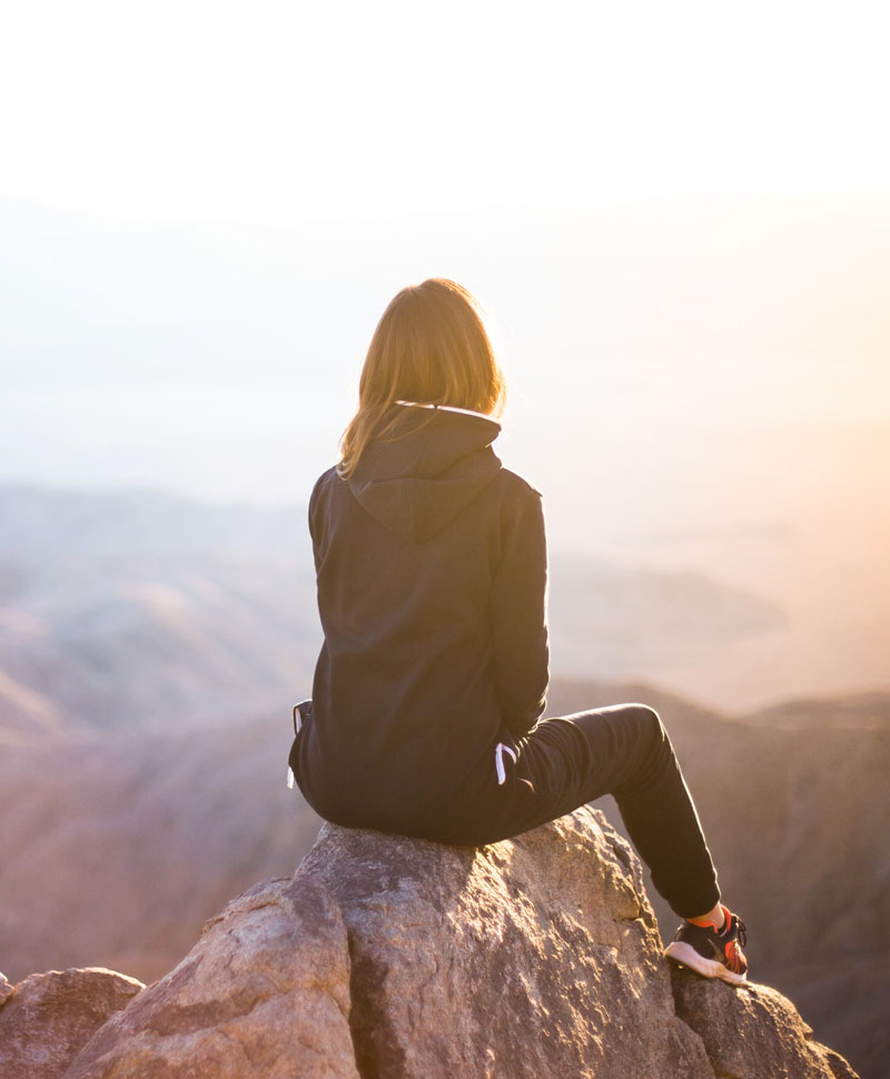 Women sitting on a rocky mountain with her back to us looking at a sunrise